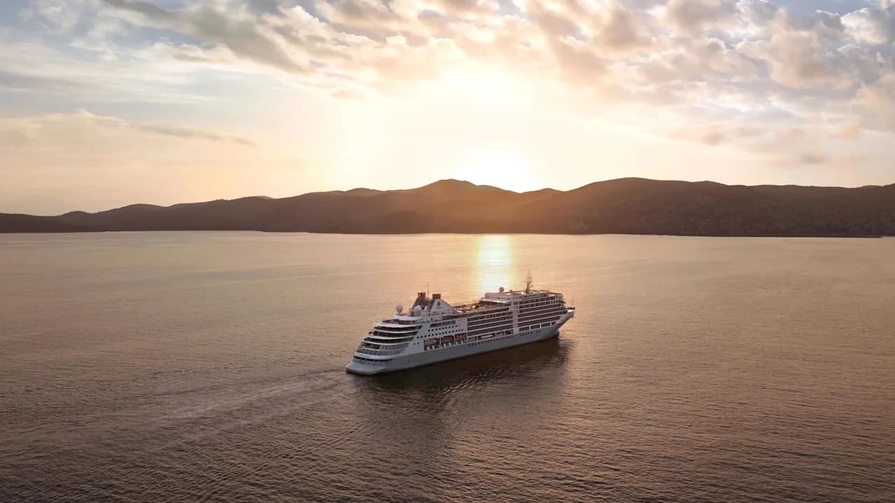 Luxury cruise ship navigating golden sunset harbor waters framing distant Kusadasi Turkey hillsides.