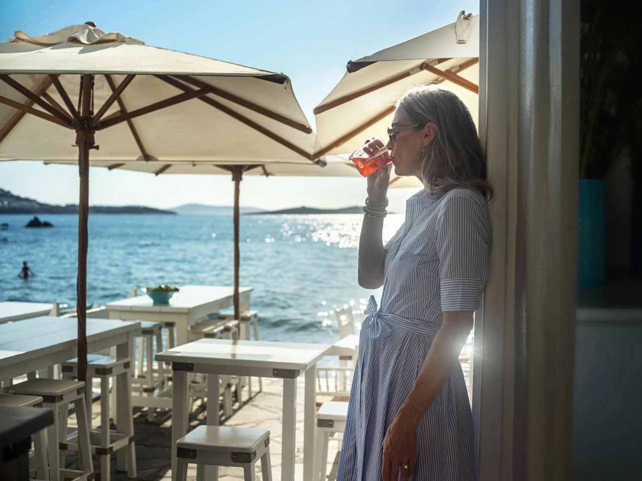 Woman drinking orange cocktail, sunny seaside Greek restaurant background.