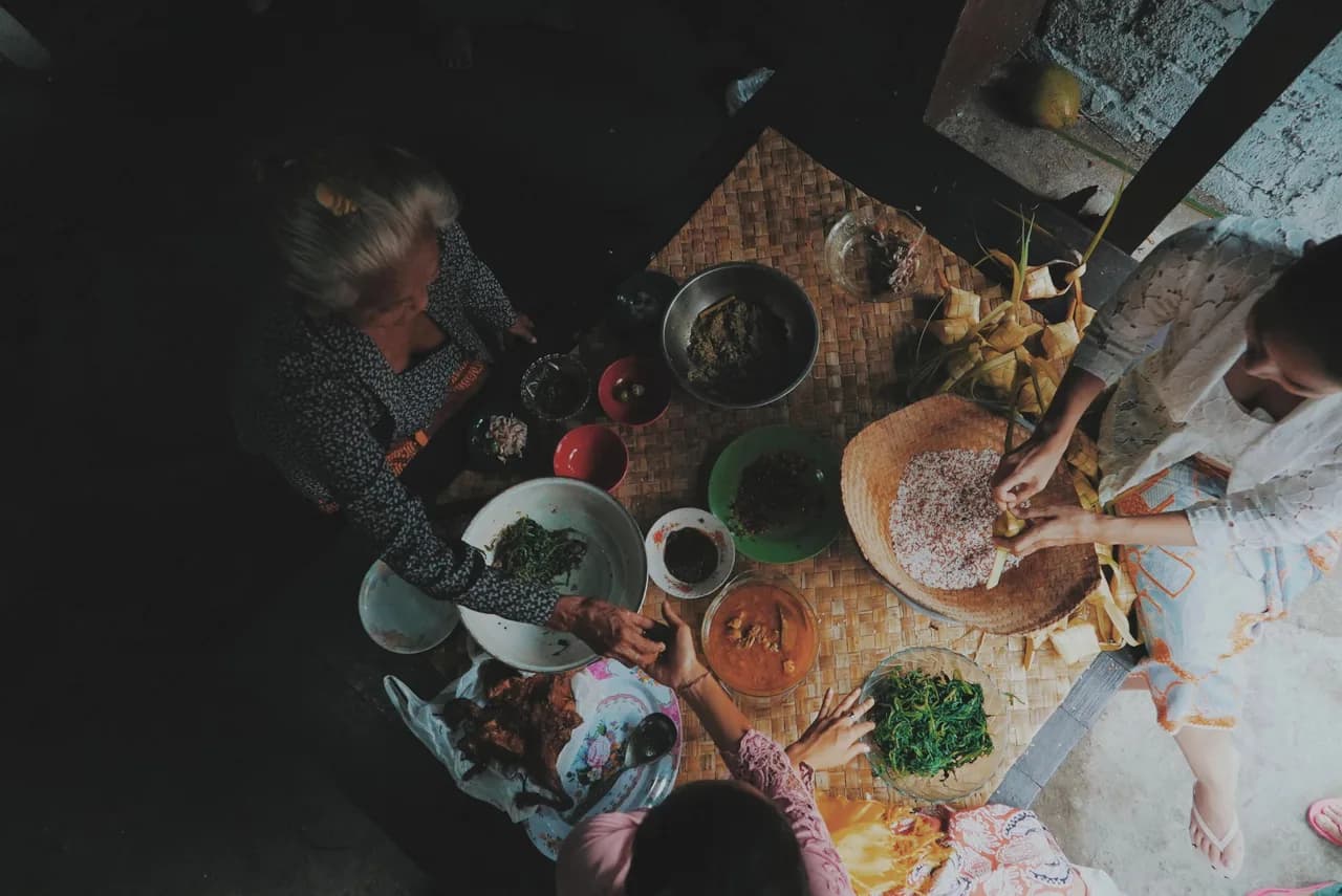 Balinese families gathering indoors to share traditional ceremonial dishes spread upon a woven bamboo mat.